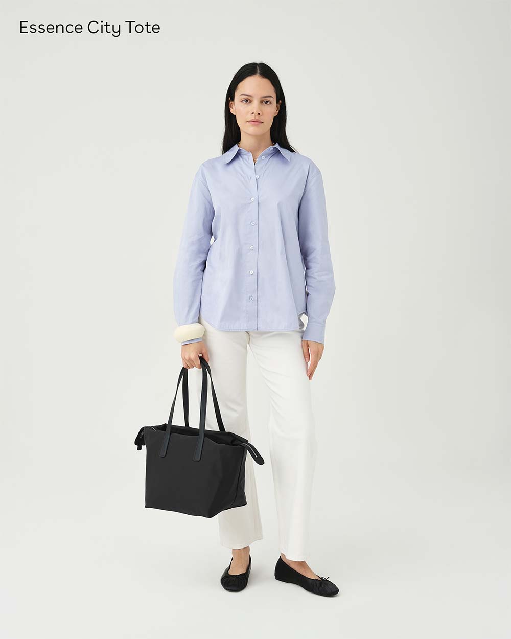 A woman stands against a light gray studio background wearing a light blue button-up shirt, white pants, and black flats, while holding an Essence City Tote in Black. The text 'Essence City Tote' appears in the top left corner.