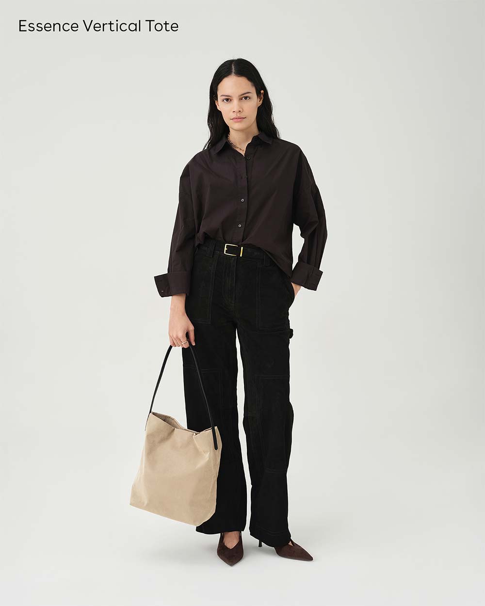 A model stands against a white studio background wearing a dark brown button-up shirt, black wide-leg pants, and brown pointed heels, while holding an Essence Vertical Tote in beige. The text 'Essence Vertical Tote' appears in the top left corner