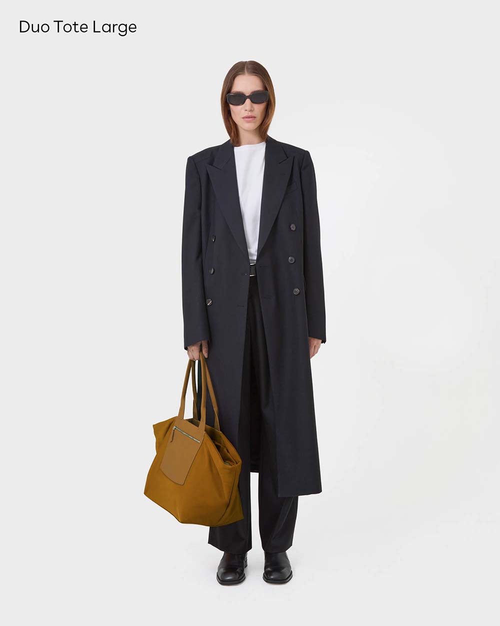 A model stands against a white studio background wearing a long black double-breasted coat over a white top and black trousers, holding a large essence duo.  Text in the top left reads 'Duo Tote Large'.