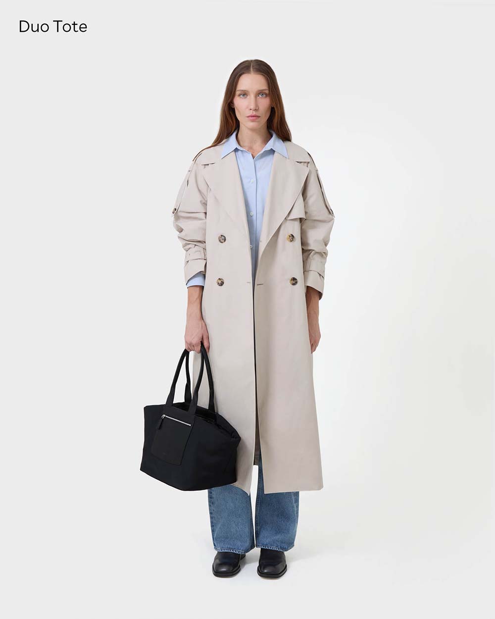 A model stands against a white studio background wearing a long beige trench coat over a light blue collared shirt, blue jeans, and black shoes, while holding Duo black tote bag. The text 'Duo Tote' appears in the top-left corner.