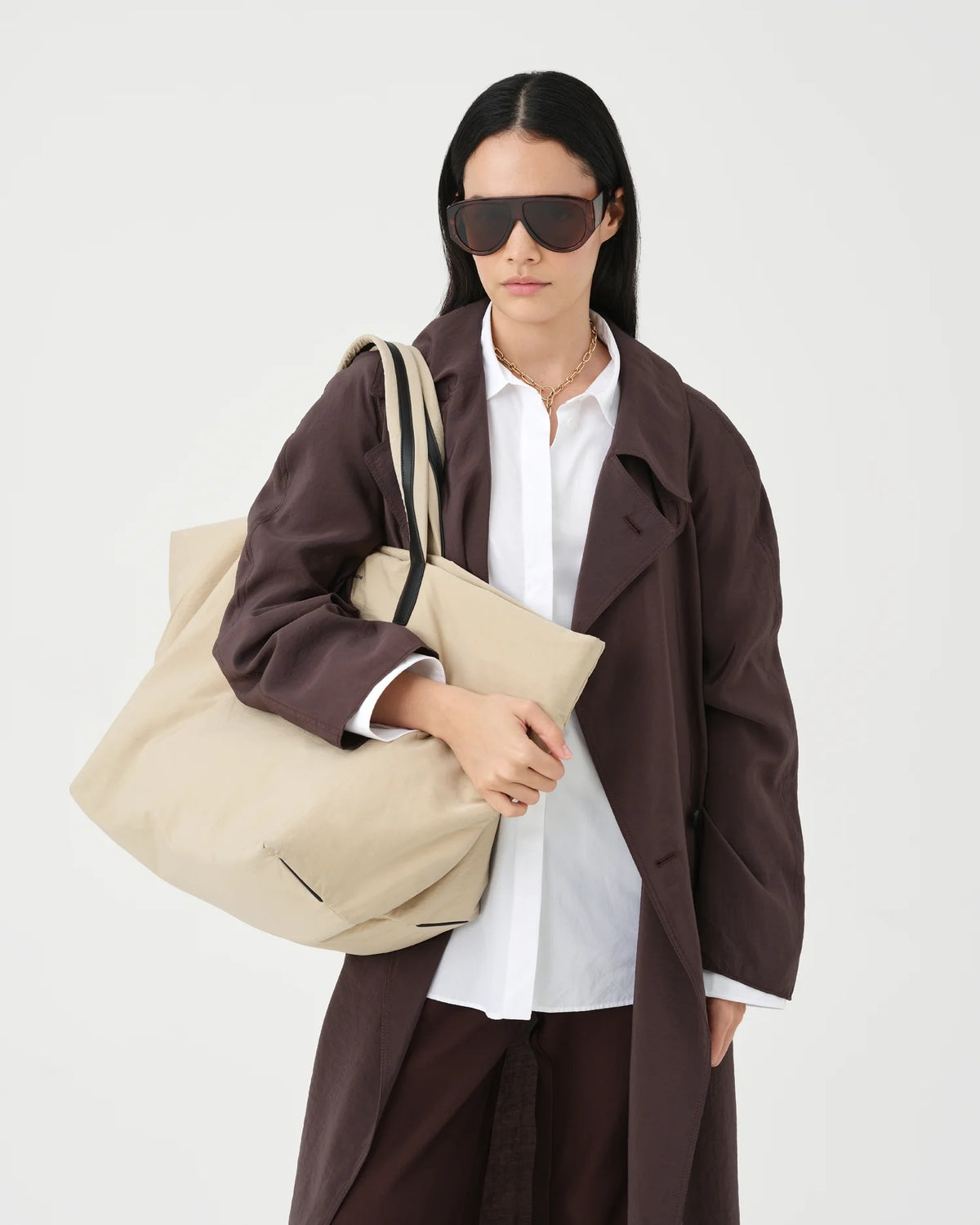 A photo of a person wearing a brown coat and holding a beige Essence Tote Bag against a white background.