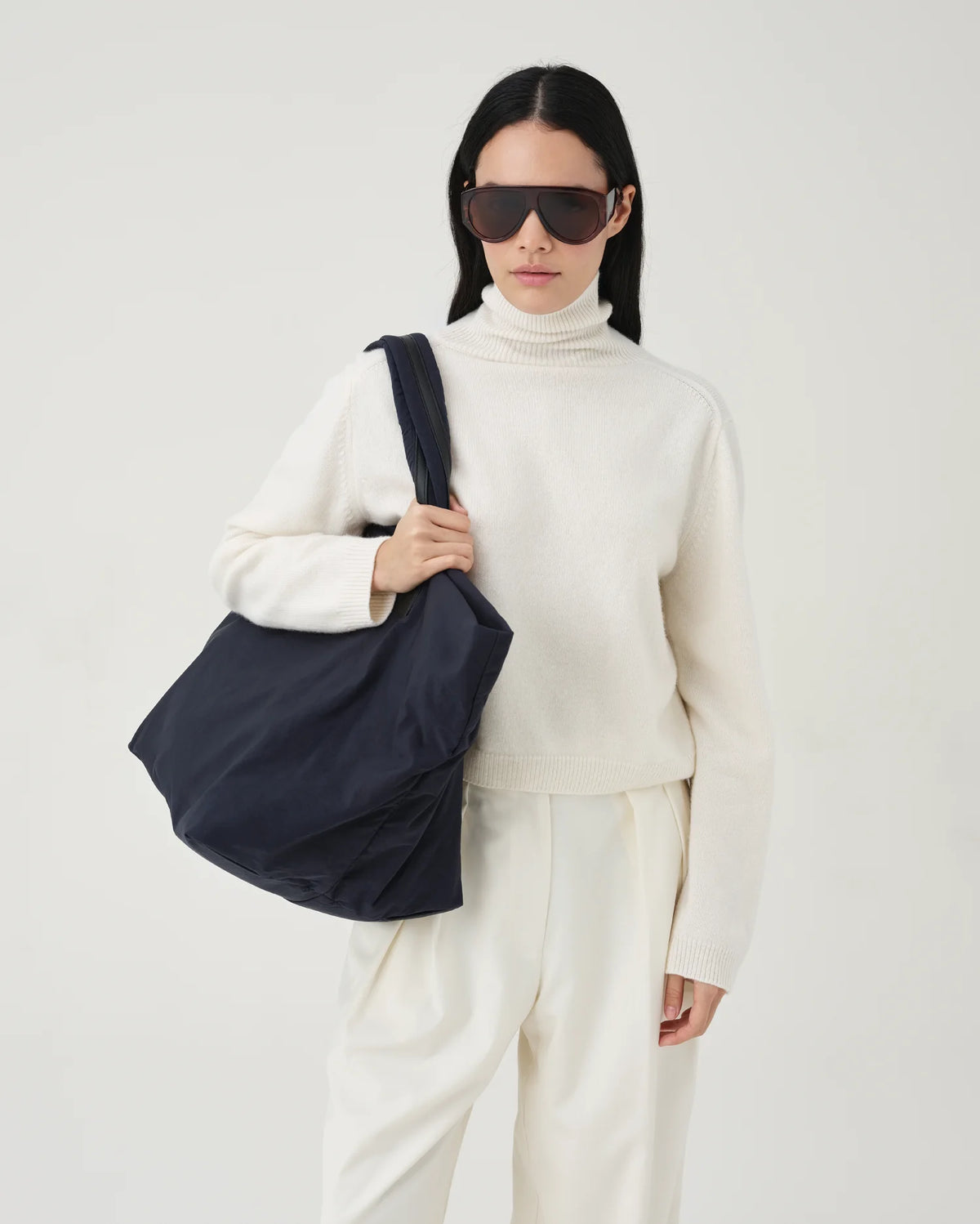 A photo of a model wearing a beige sweater, white pants, and sunglasses, holding a quilted Essence Tote Bag against a white background.
