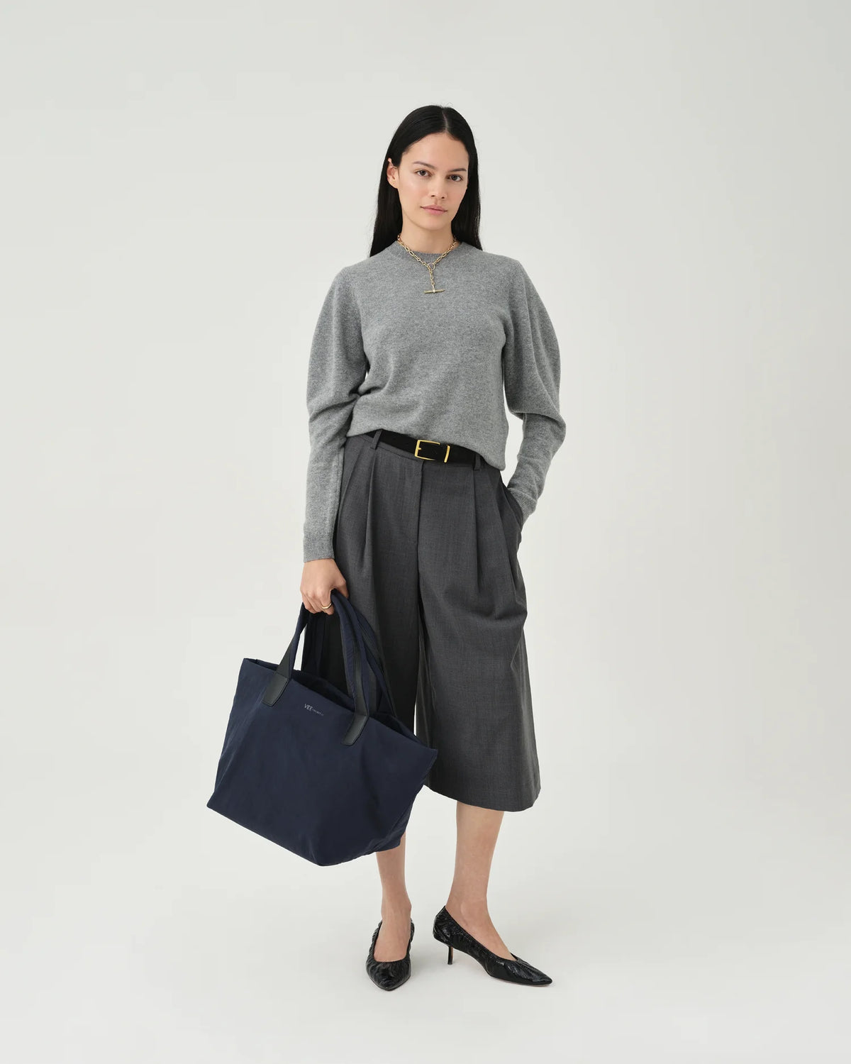 A photo of a model wearing a beige sweater, white pants, and sunglasses, holding a quilted Essence Tote Bag against a white background.