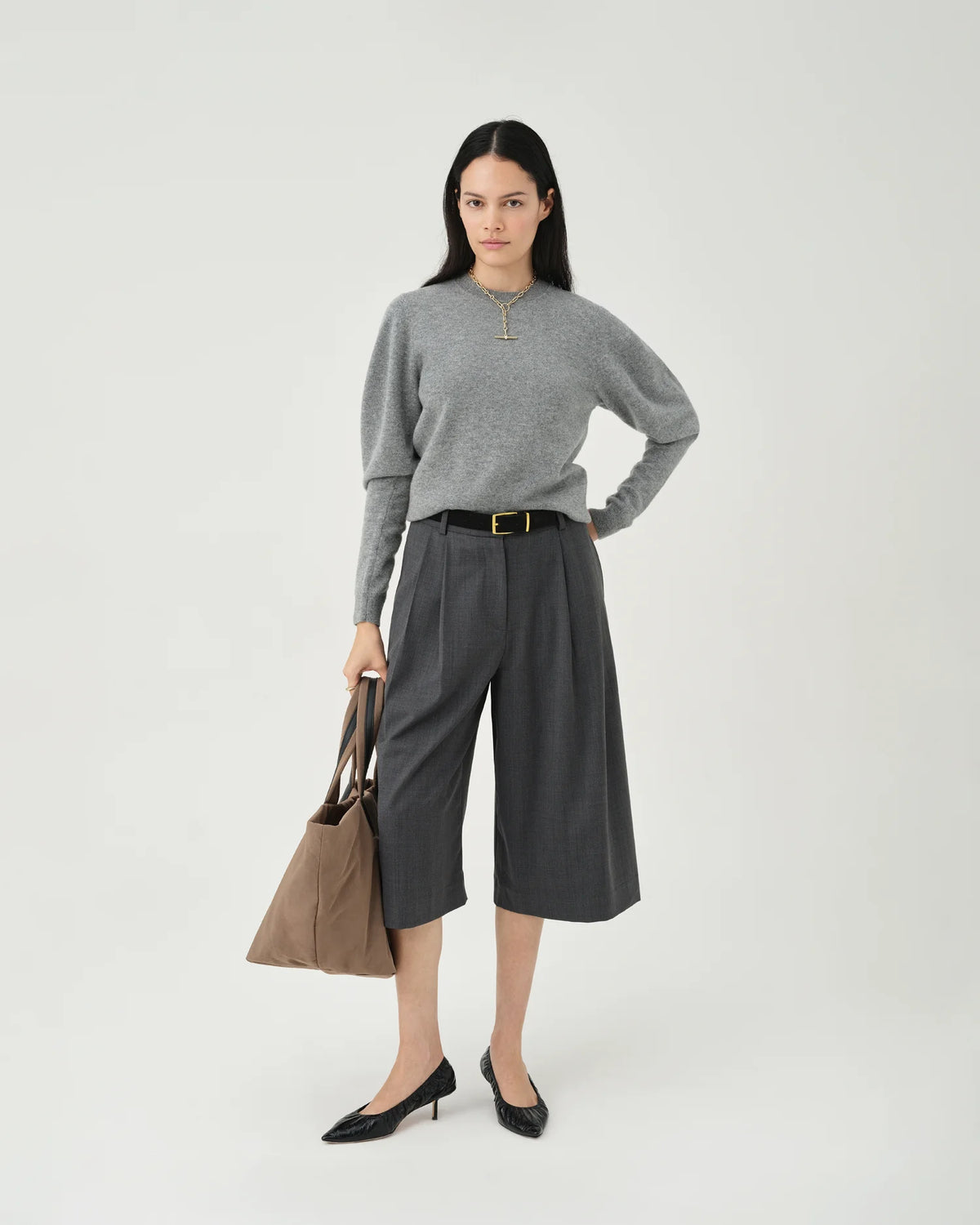 A photo of a model wearing a gray sweater and dark pants, holding a mink Essence Tote Bag against a white background.