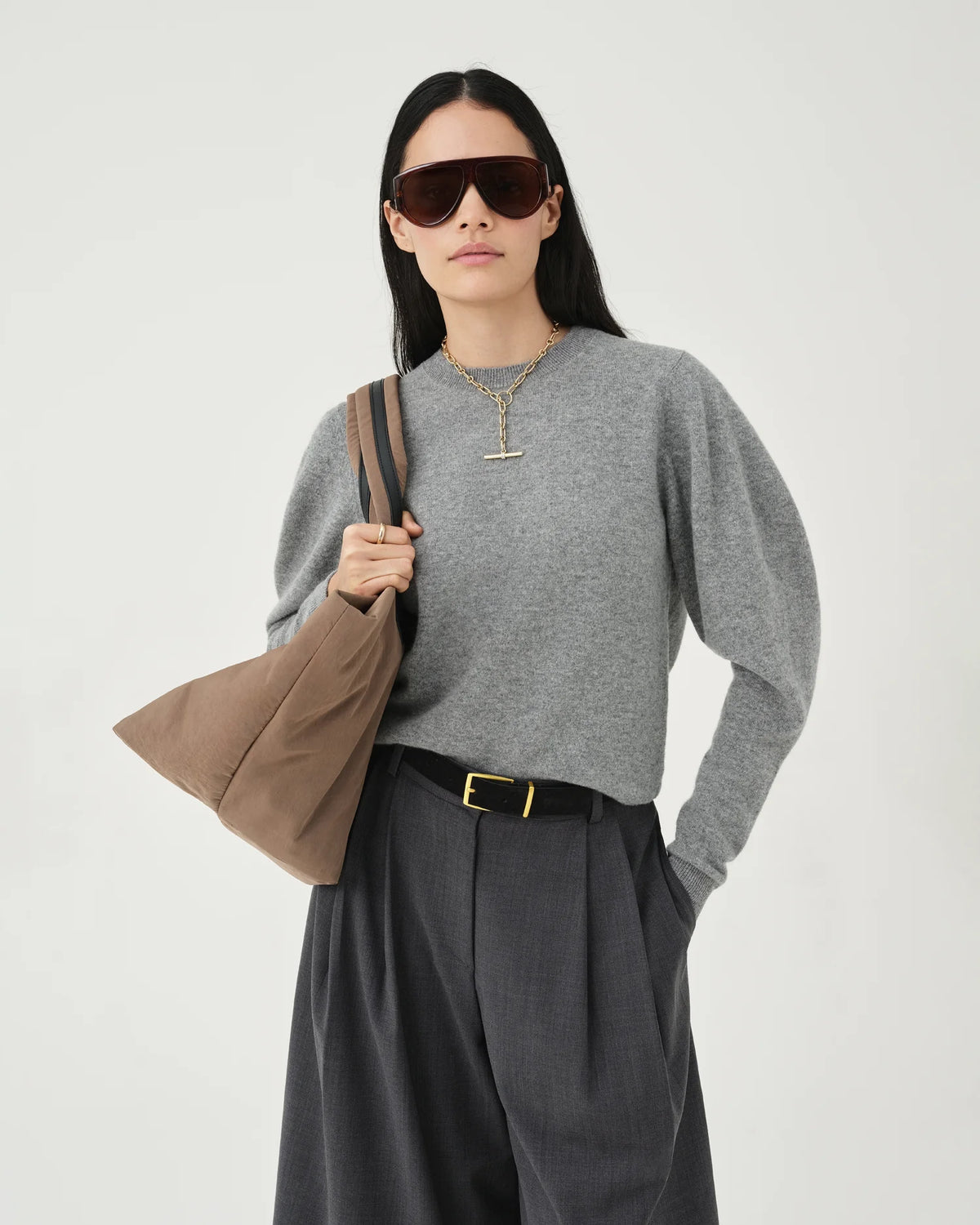 A photo of a model wearing a gray sweater and dark pants, holding a mink Essence Tote Bag against a white background.