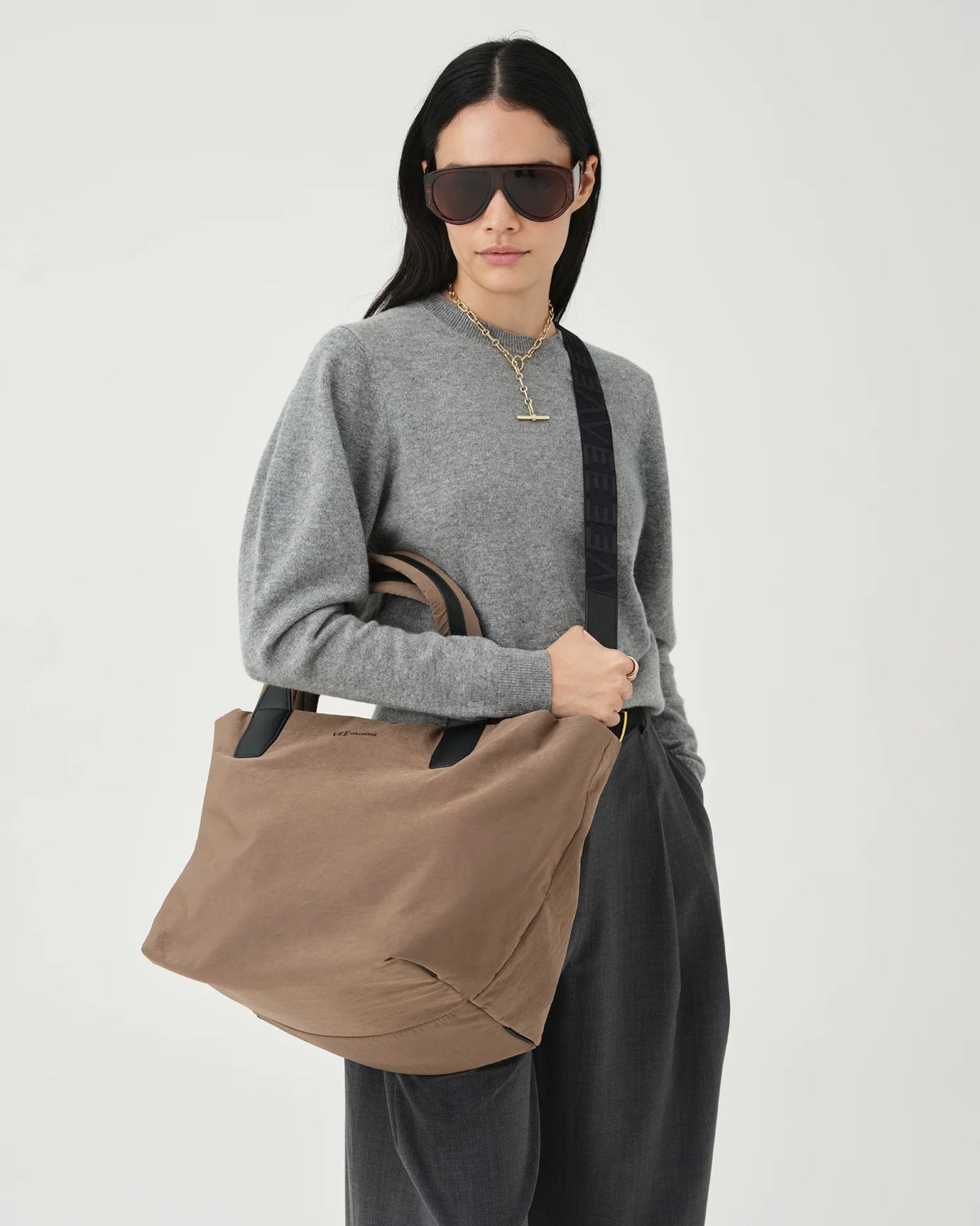 A photo of a model wearing a gray sweater and dark pants, holding a mink Essence Tote Bag against a white background.