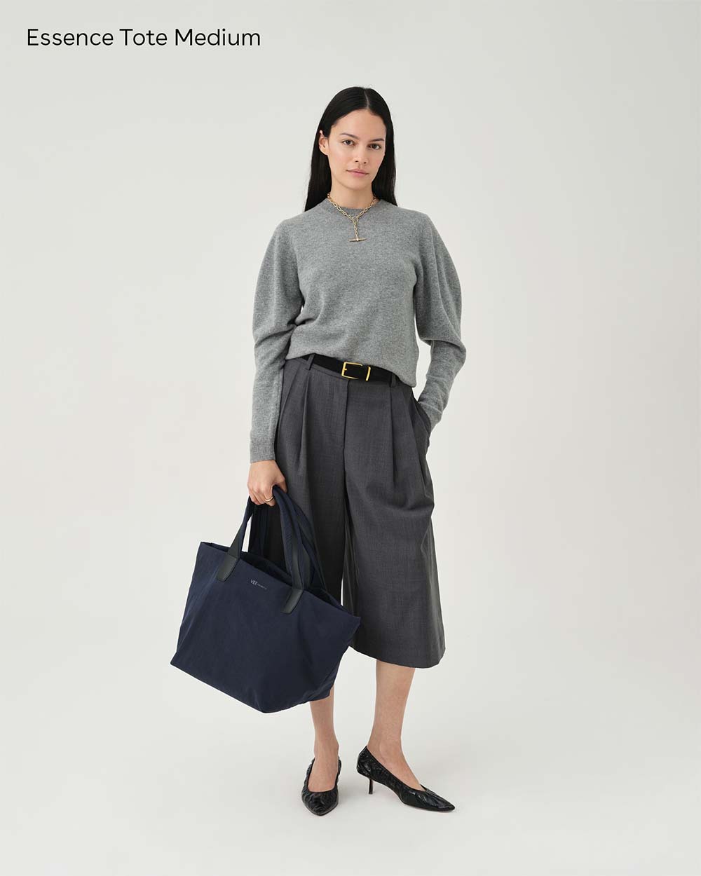 A model stands against a white studio background wearing a gray knit sweater, dark gray pleated wide-leg pants, black pointed heels, and holding Essence Tote Medium Midnight Trench. Text in the top-left reads 'Essence Tote Medium'.