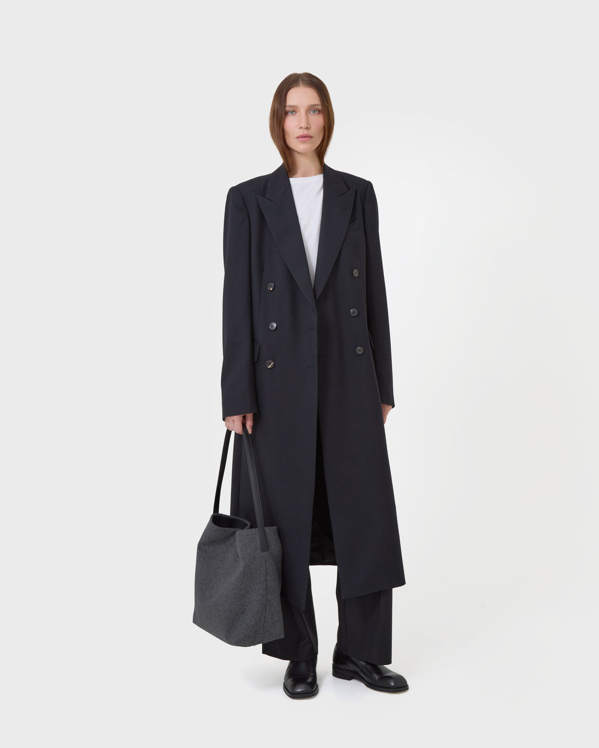 A model stands against a light gray studio background wearing a long black double-breasted overcoat over a white top and black trousers, while holding a dark gray tote bag.