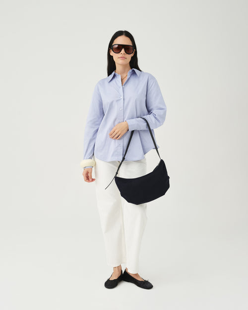 A model wearing a light blue button-up shirt, white pants, and dark sunglasses holds an Essence Wool Banana bag with black handles against a white studio background.