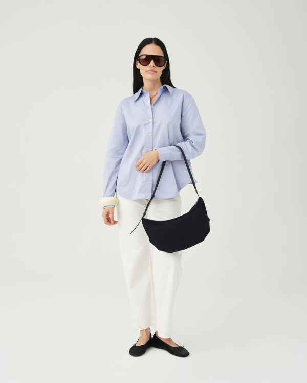 A model wearing a light blue button-up shirt, white pants, and dark sunglasses holds an Essence Wool Banana bag with black handles against a white studio background.