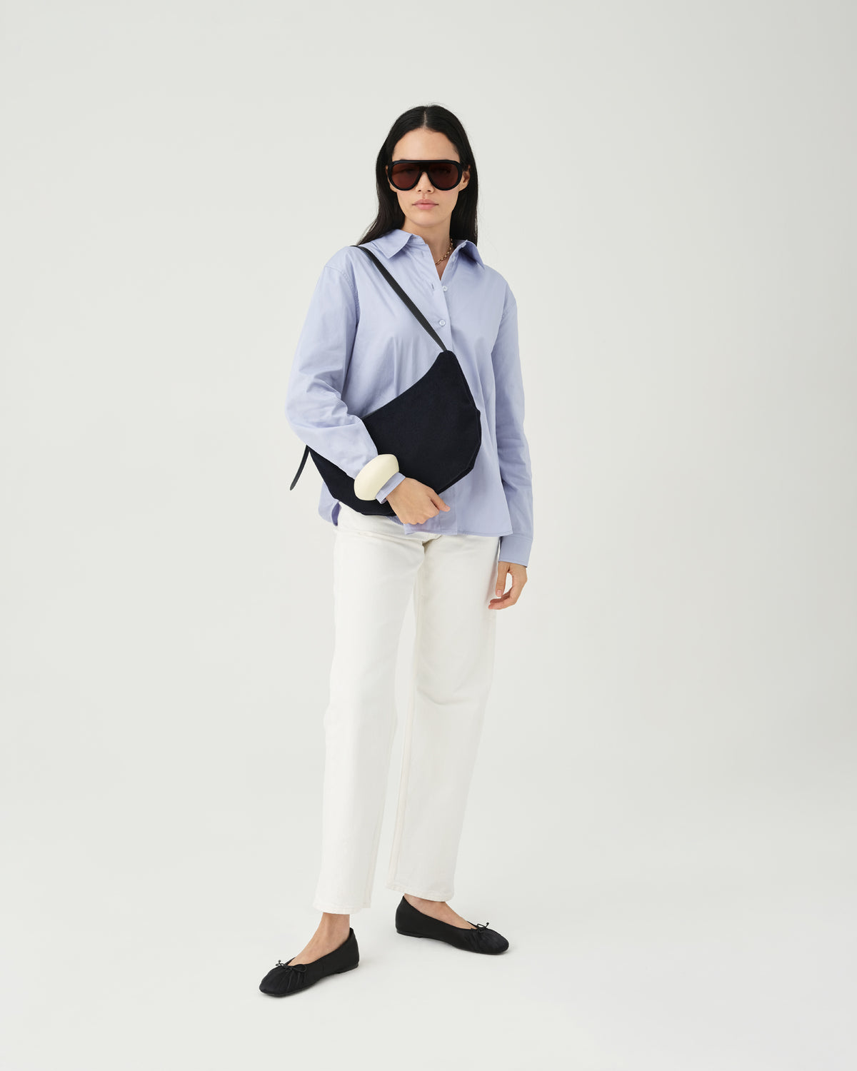 A model wearing a light blue button-up shirt, white pants, and dark sunglasses holds an Essence Wool Banana bag with black handles against a white studio background.