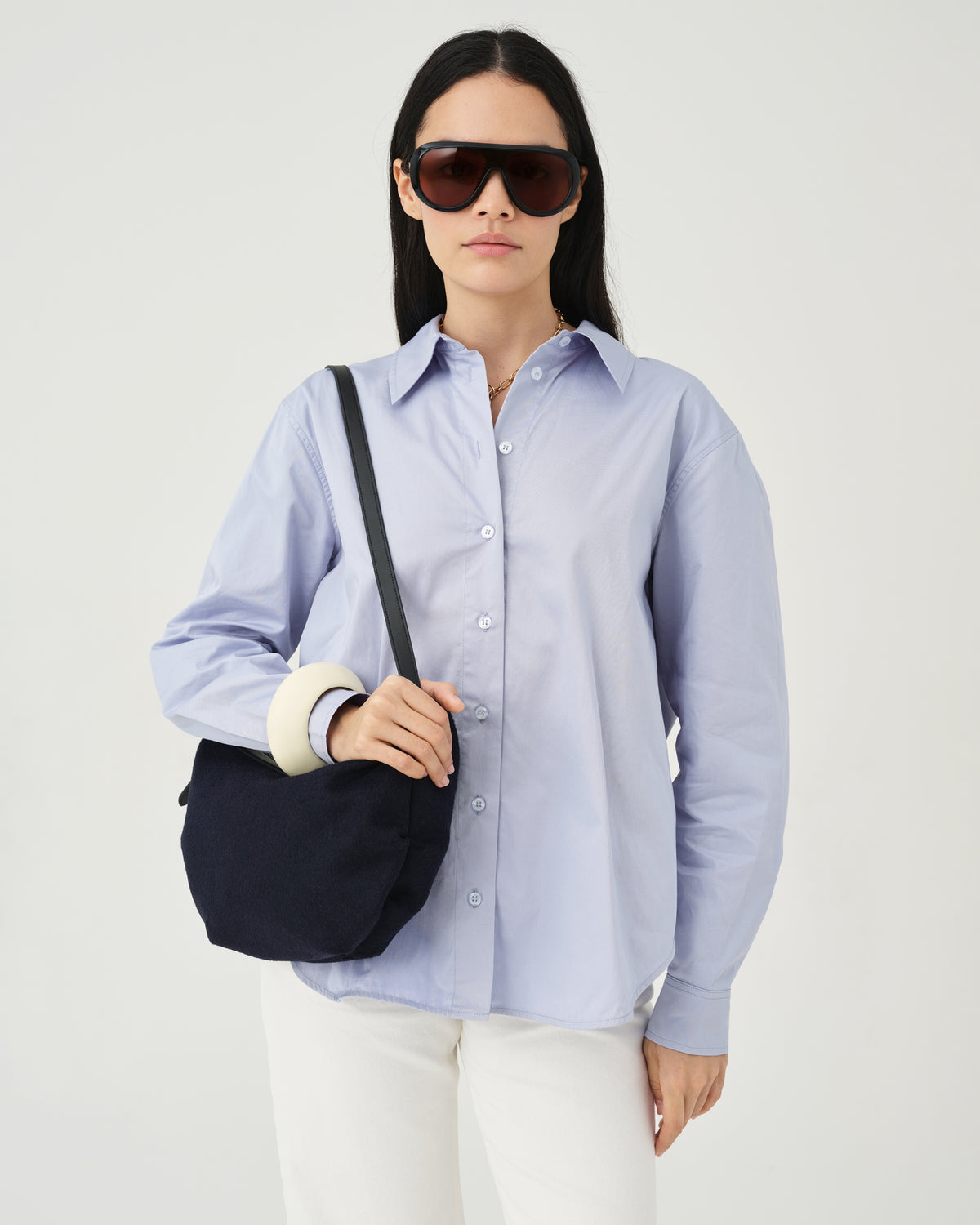 A model wearing a light blue button-up shirt, white pants, and dark sunglasses holds an Essence Wool Banana bag navy with black handles against a white studio background.