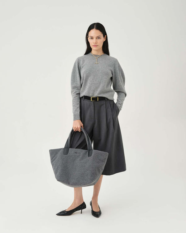 A model stands against a light gray studio background wearing a gray mock-neck knit sweater, dark gray pleated midi skirt with a gold buckle, black pointed-toe heels, and holding a large gray fabric tote bag. c__2