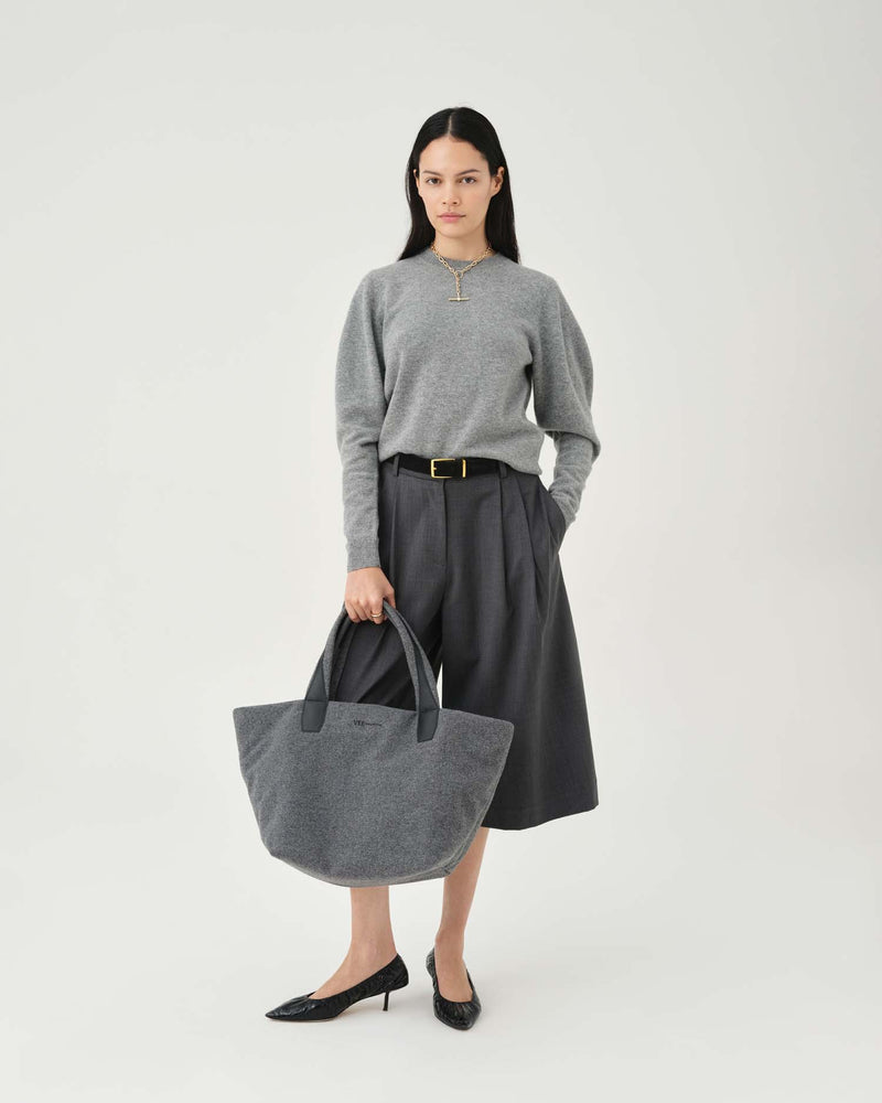 A model stands against a light gray studio background wearing a gray mock-neck knit sweater, dark gray pleated midi skirt with a gold buckle, black pointed-toe heels, and holding a large gray fabric tote bag.
