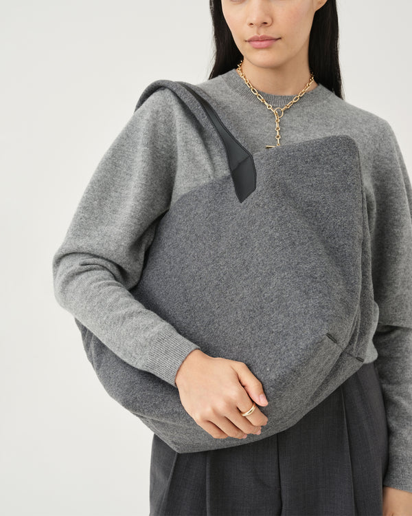 A studio fashion photo of a woman wearing a heather gray long-sleeve knit top with dramatic balloon sleeves, paired with dark gray wide-leg trousers and a black belt with a gold buckle, carrying an Essence Wool Tote bag against a clean white background.