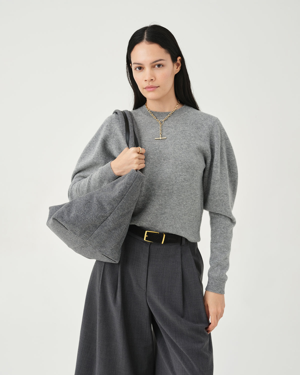 A studio fashion photo of a woman wearing a heather gray long-sleeve knit top with dramatic balloon sleeves, paired with dark gray wide-leg trousers and a black belt with a gold buckle, carrying an Essence Wool Tote bag against a clean white background.