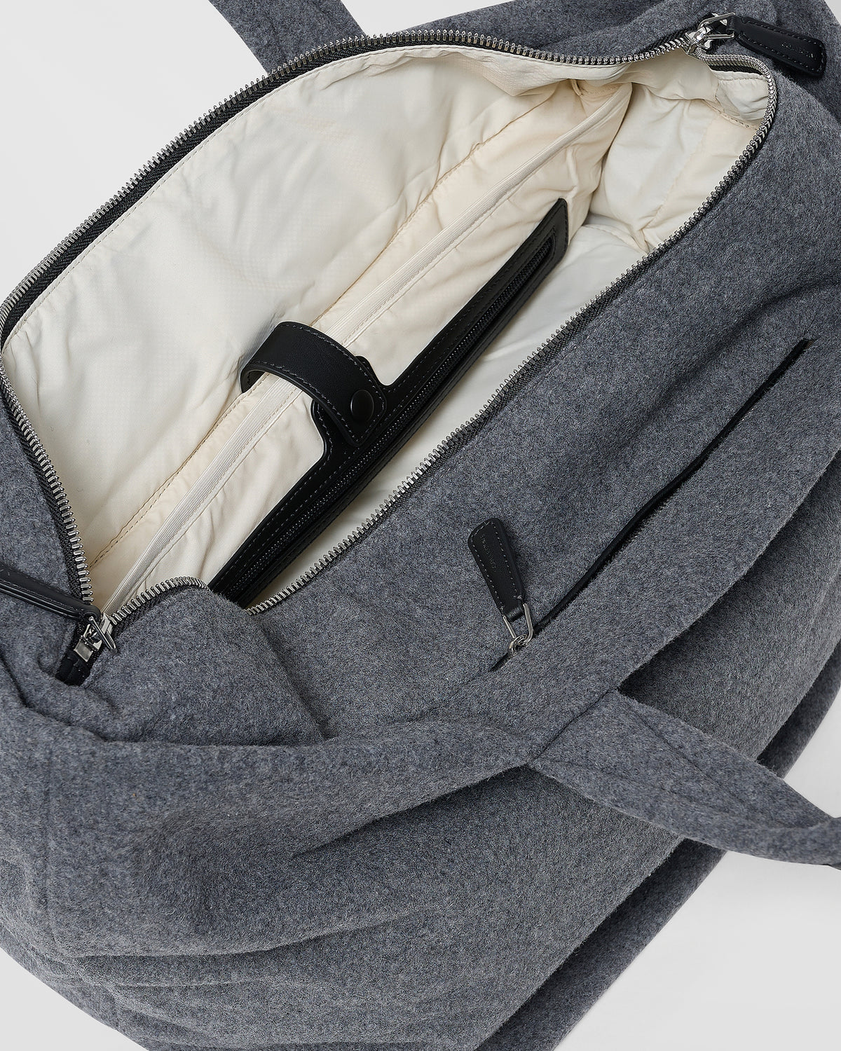A close-up product photo of an open Essence Wool Tote bag, showcasing its cream-colored interior lining, black straps, and silver zipper hardware.