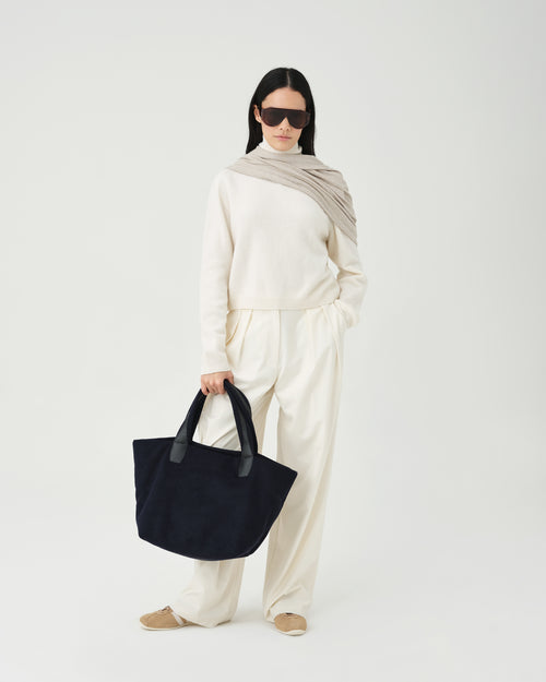 A studio fashion photo of a model wearing a cream knit sweater and trousers, a beige shawl, and dark sunglasses, carrying an Essence Wool Tote bag navy over her shoulder against a clean white background.