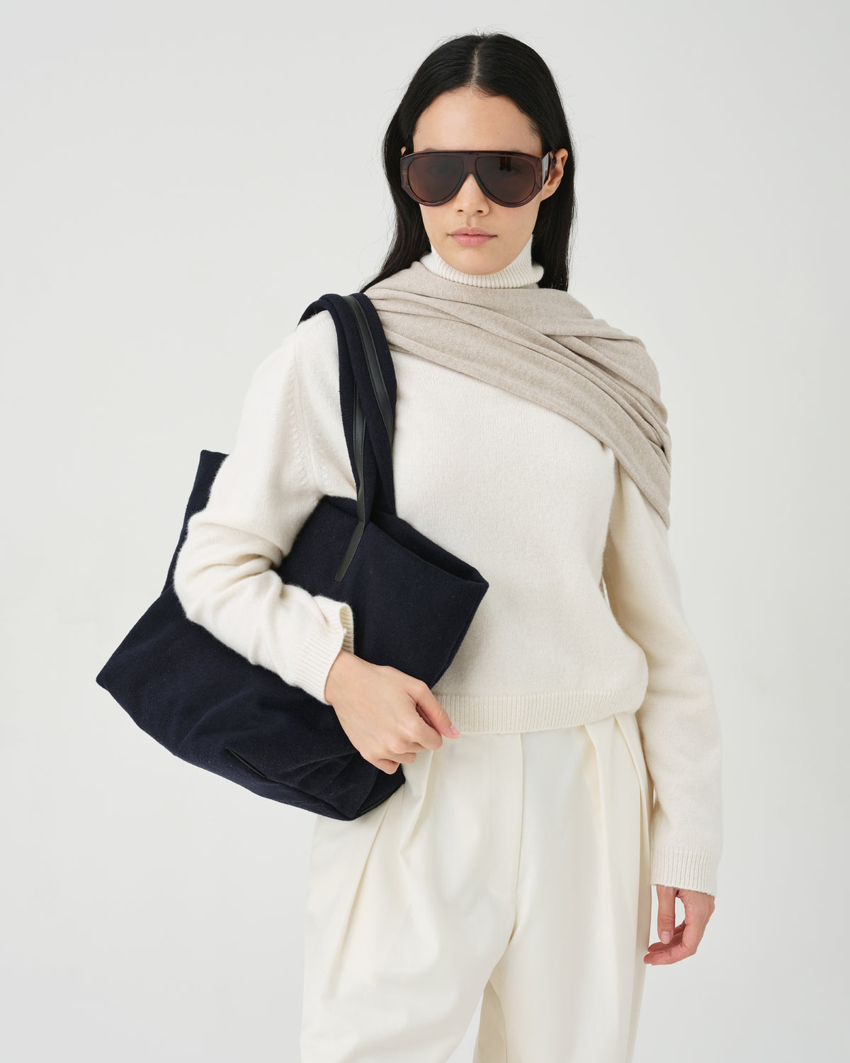 A studio fashion photo of a model wearing a cream knit sweater and trousers, a beige shawl, and dark sunglasses, carrying an Essence Wool Tote bag over her shoulder against a clean white background.