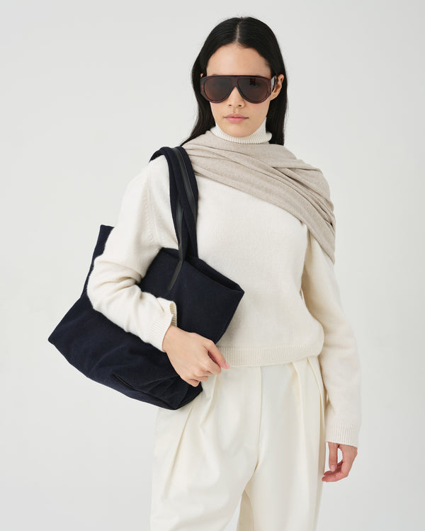 A studio fashion photo of a model wearing a cream knit sweater and trousers, a beige shawl, and dark sunglasses, carrying an Essence Wool Tote bag over her shoulder against a clean white background.