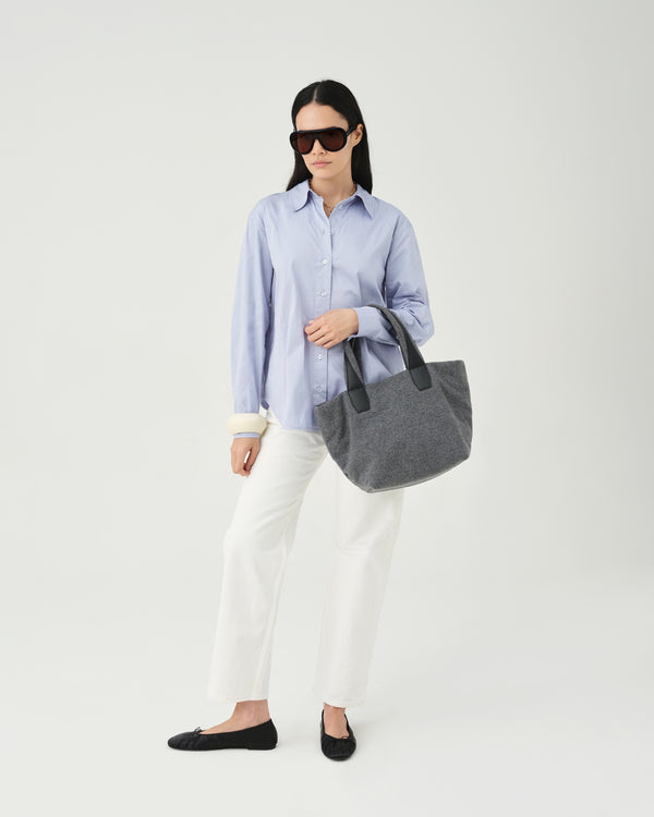 A model wearing a light blue button-up shirt, white pants, and dark sunglasses holds an Essence Wool Tote bag with black handles against a white studio background.