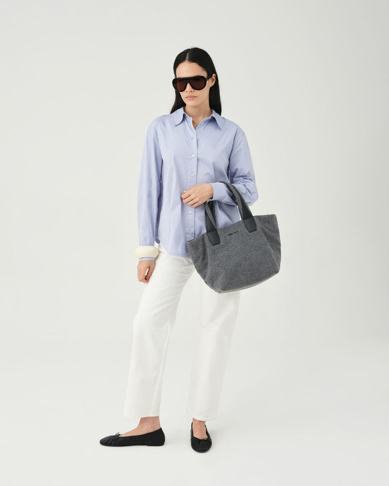 A woman modeling a light blue striped button-down shirt, white trousers, black flats, and sunglasses while carrying a large gray fabric tote bag against a plain light gray studio background.