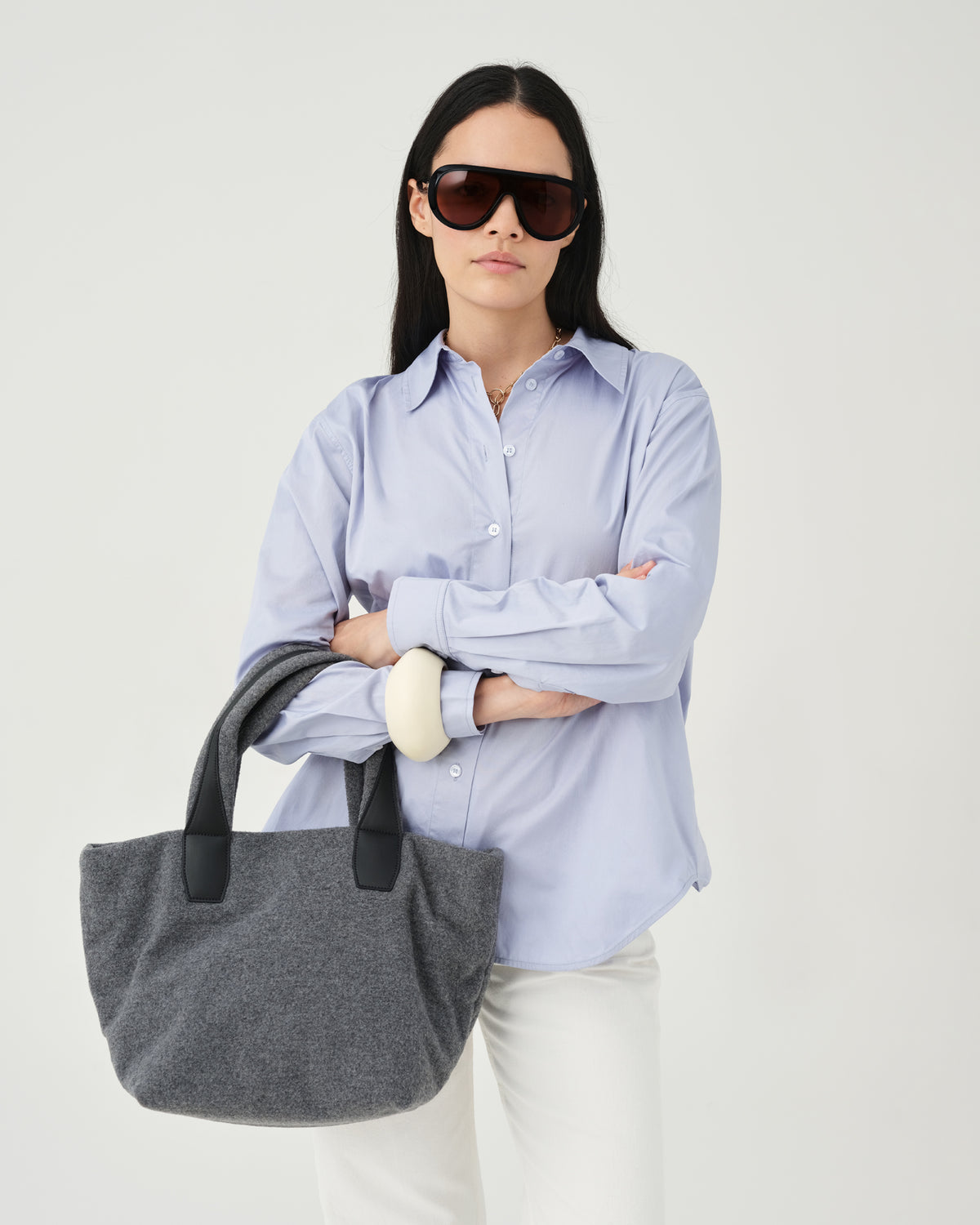 A model wearing a light blue button-up shirt, white pants, and dark sunglasses holds an Essence Wool Tote bag with black handles against a white studio background.