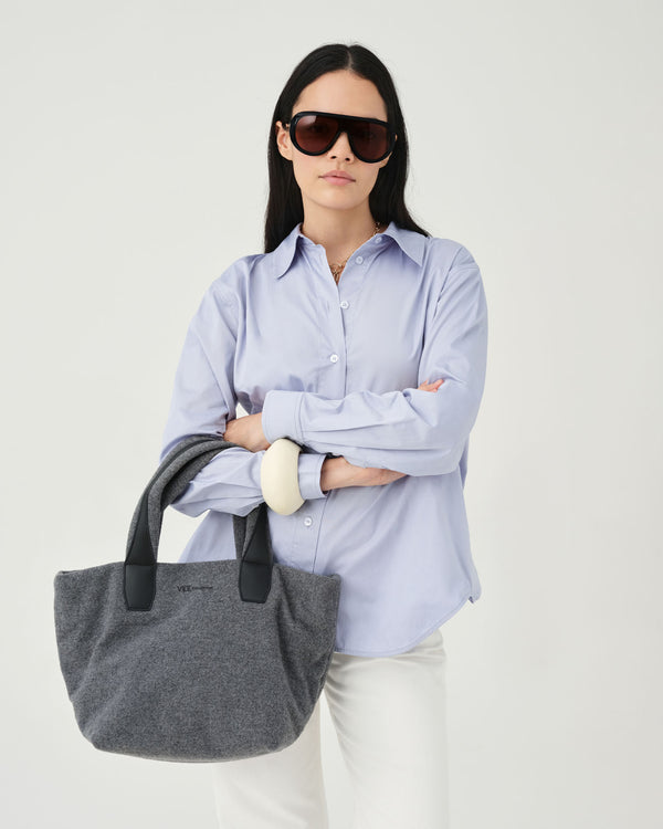 A woman wearing a light blue button-up shirt, white pants, and dark sunglasses holds a large gray fabric tote bag with black handles against a plain light background.