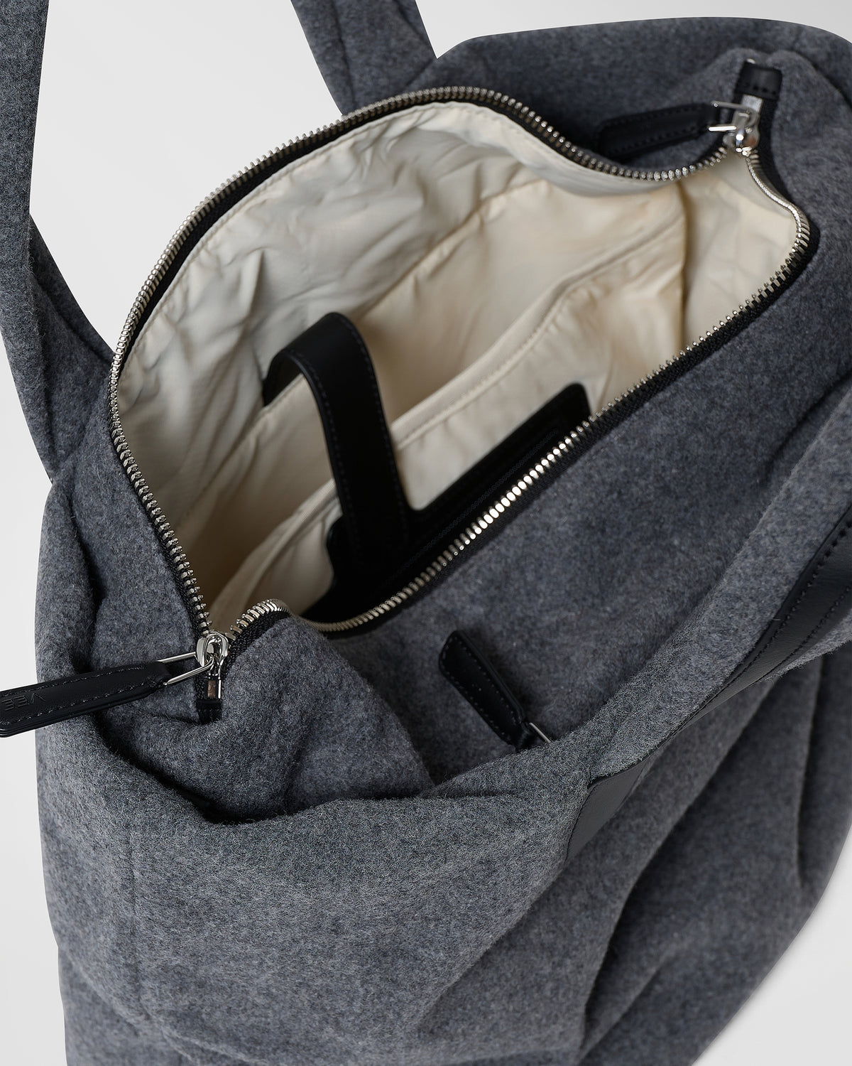 A close-up product photo of an open Essence Wool Tote bag, showcasing its cream-colored interior lining, black straps, and silver zipper hardware.