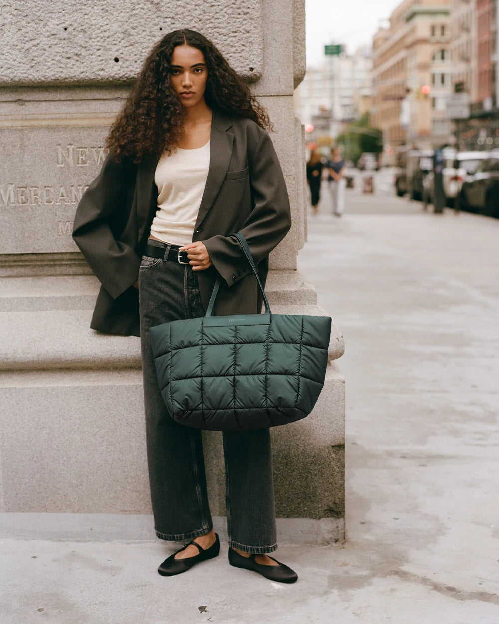 A woman standing on a city sidewalk wearing a dark oversized blazer, white tank top, and black jeans, holding a large quilted Vee Collective Porter Marche bag in bottle green by her side.