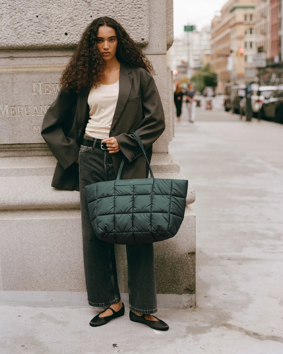A woman standing on a city sidewalk wearing a dark oversized blazer, white tank top, and black jeans, holding a large quilted Vee Collective Porter Marche bag in bottle green by her side.