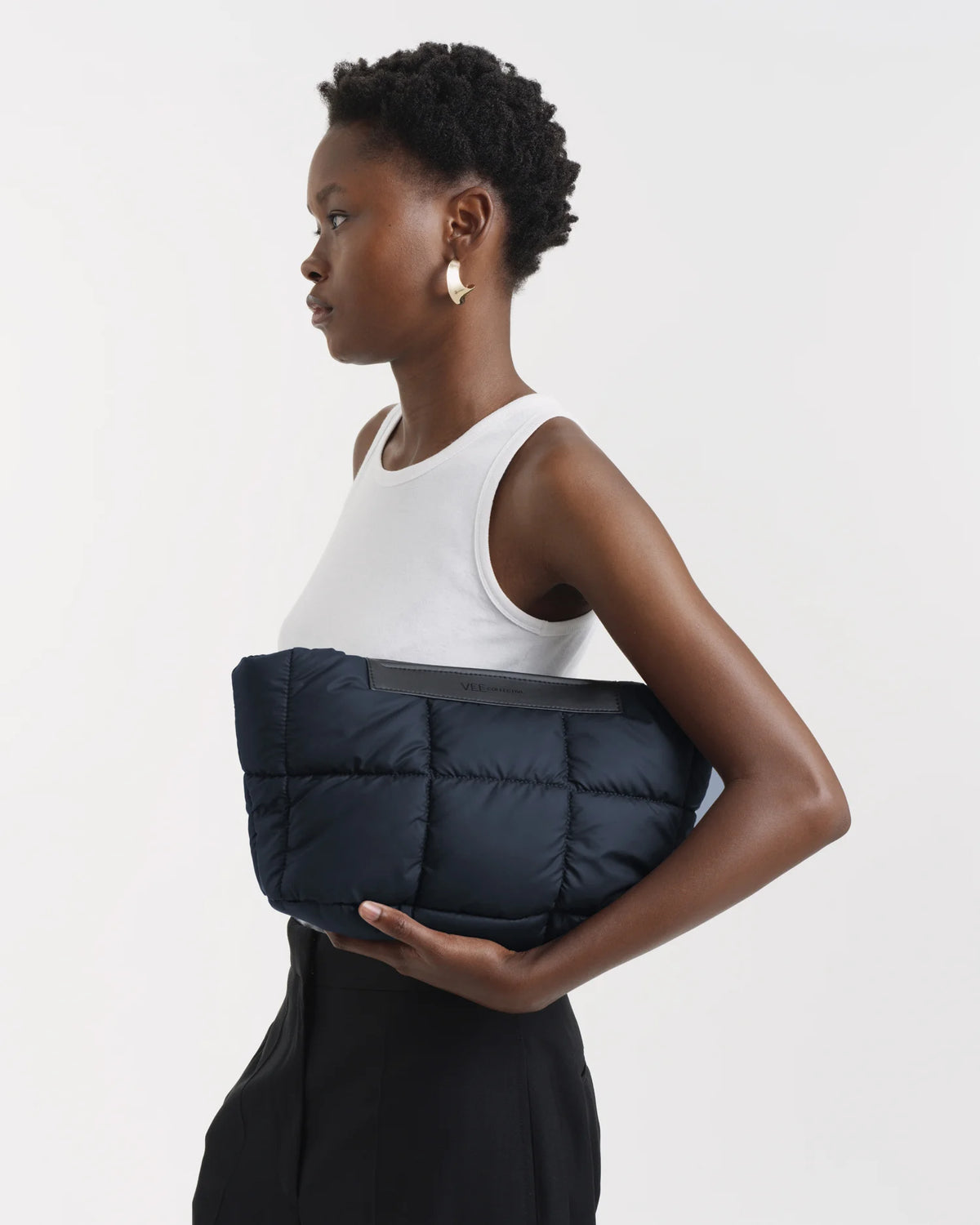 On-body picture of a woman holding the Porter Basque Clutch Bag in Dark Navy by Vee Collective, wearing a minimalistic outfit against a clean studio background.