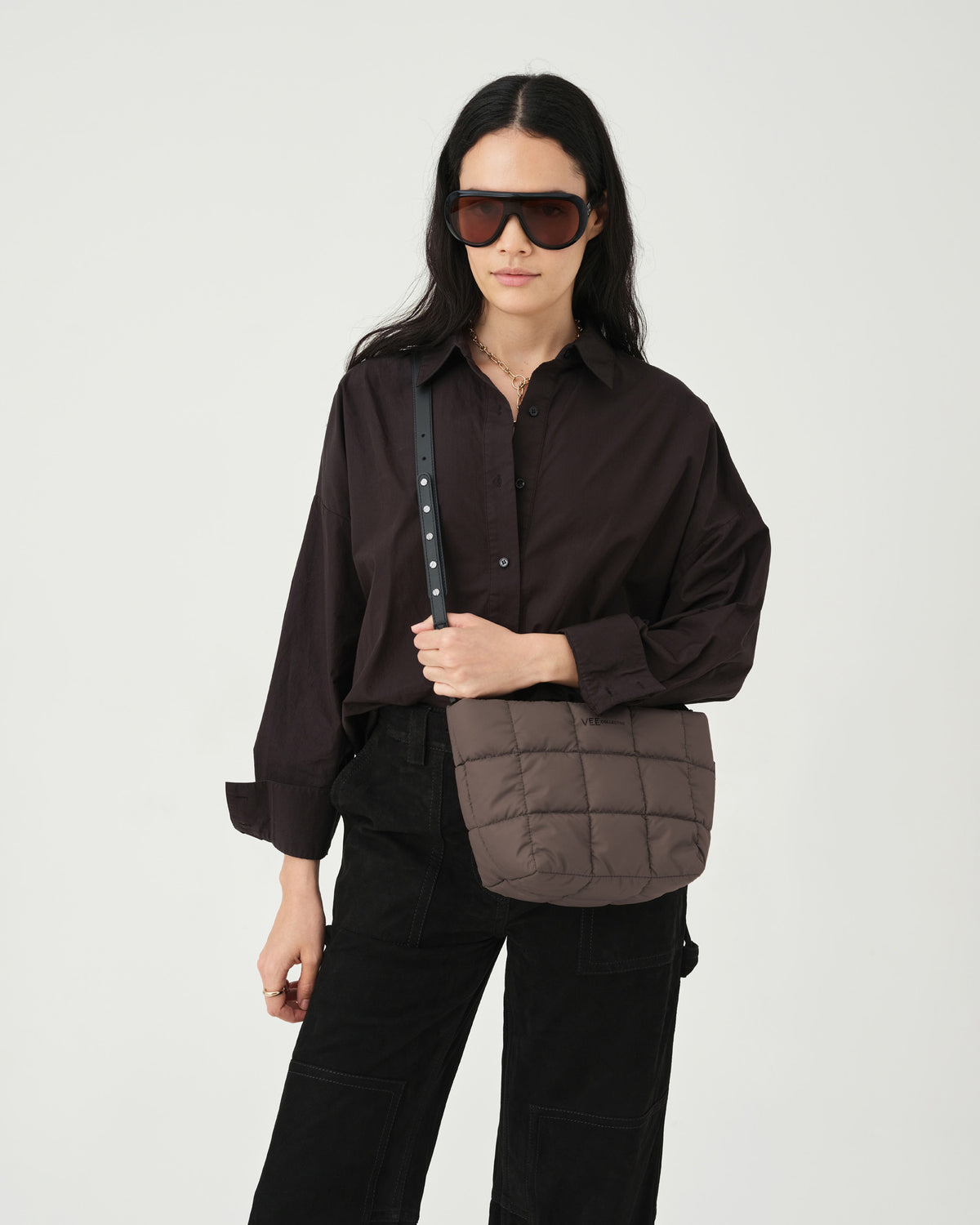 A model stands against a light backdrop, holding a small quilted tote bag in a soft brown color. She wears a dark button-up shirt and black trousers, showcasing the Porter totes mink square pattern and comfortable shoulder straps.