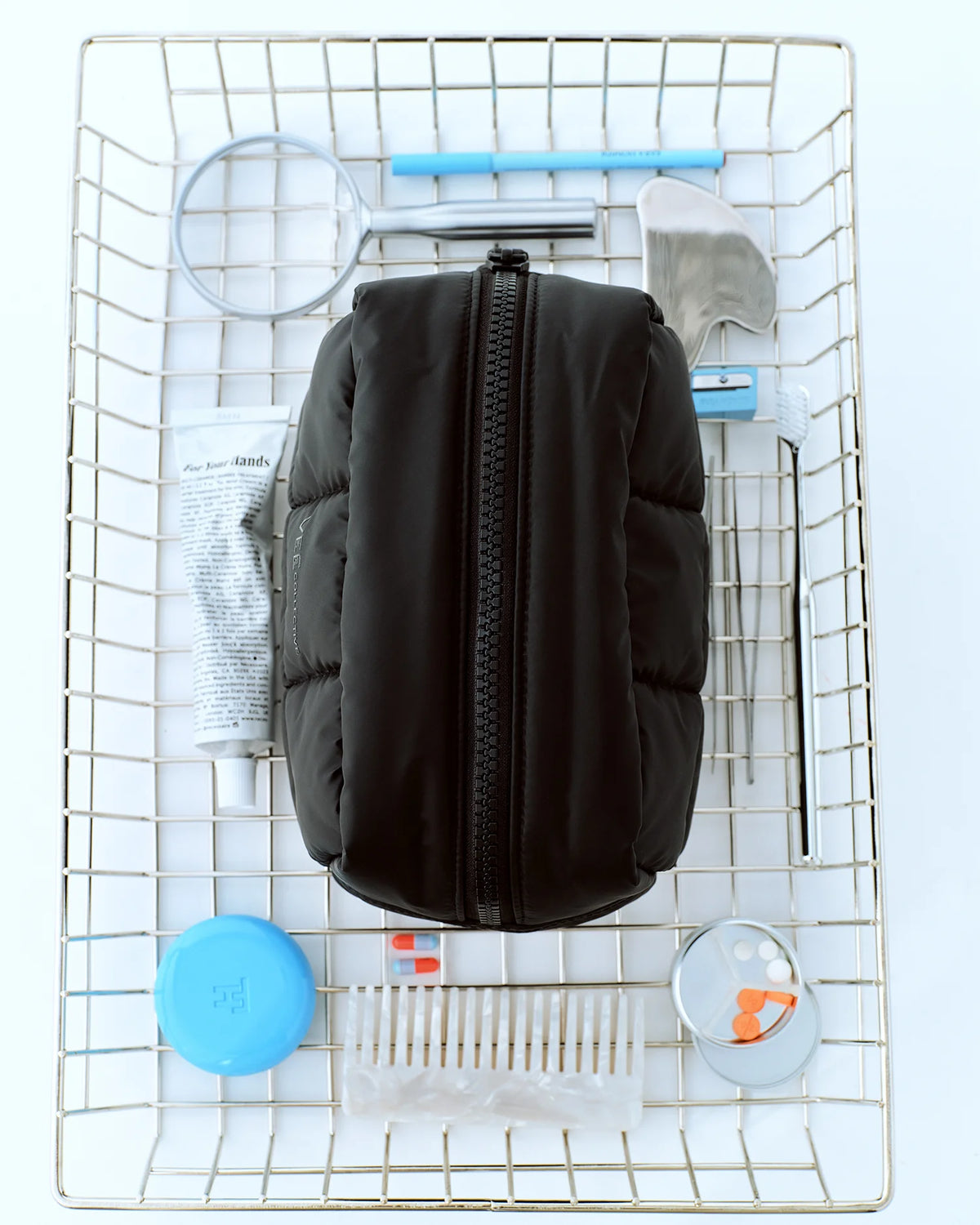 A Matt Black Porter Cosmetics Pouch Bag in Medium by Vee Collective, placed in the center of a metal wire basket, surrounded by grooming and travel essentials.