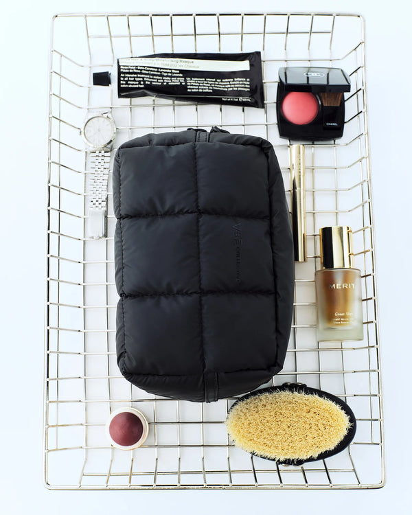 A Matt Black quilted Porter cosmetics pouch by Vee Collective placed in the center of a metal wire basket, surrounded by beauty items.