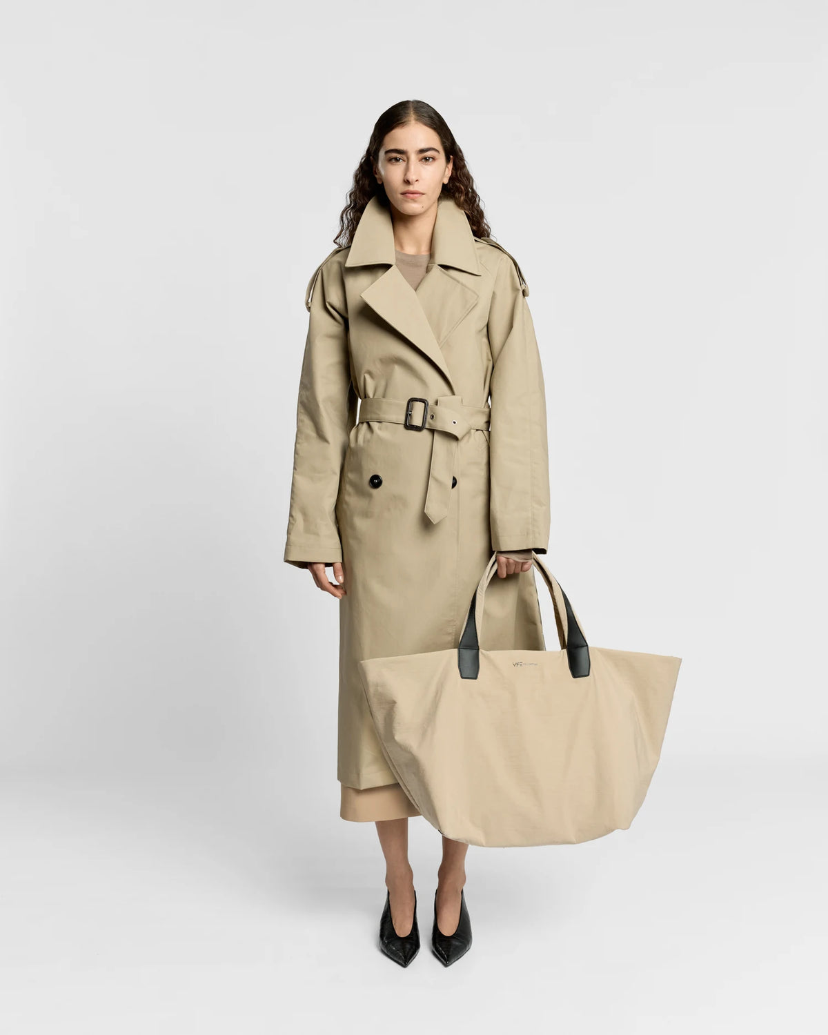 Woman in a beige trench coat holding the Porter Essence Weekender Tote Bag in Beige Trench by Vee Collective against a plain studio background.
