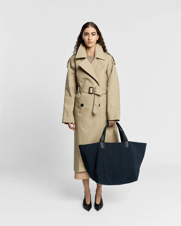 Woman in a beige trench coat holding the Porter Essence Weekender Tote Bag in Midnight Trench by Vee Collective against a plain studio background.