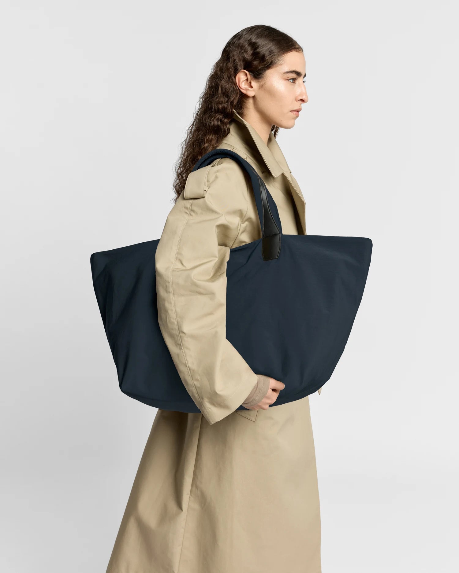 Woman in a beige trench coat carrying the Porter Essence Weekender Tote Bag in Midnight Trench by Vee Collective against a plain studio background.