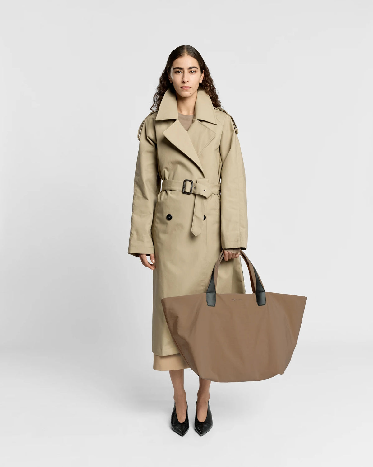 Woman in a beige trench coat holding the Porter Essence Weekender Tote Bag in Mink Trench by Vee Collective against a plain studio background.