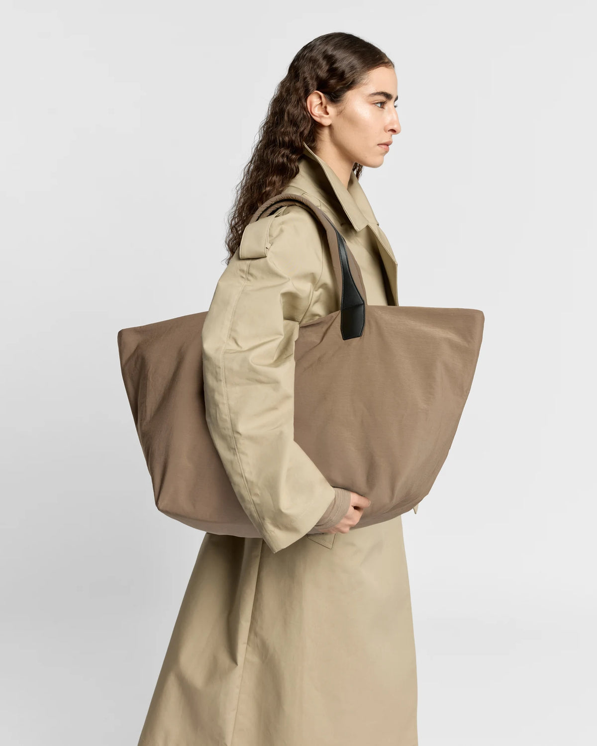 Woman in a beige trench coat carrying the Porter Essence Weekender Tote Bag in Mink Trench by Vee Collective against a plain studio background.