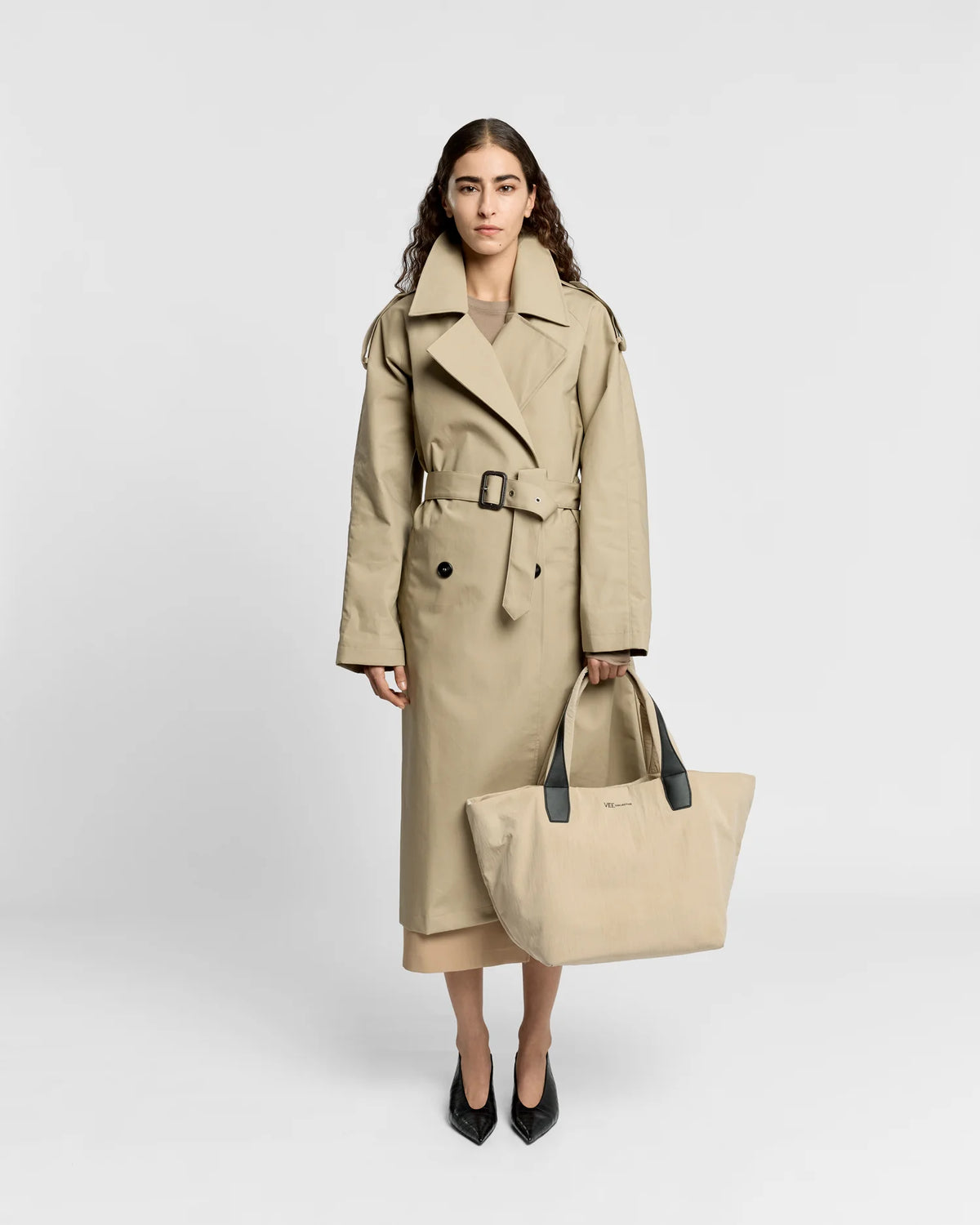 Woman in a beige trench coat holding the Porter Essence Medium Tote Bag in Beige Trench by Vee Collective against a plain studio background.