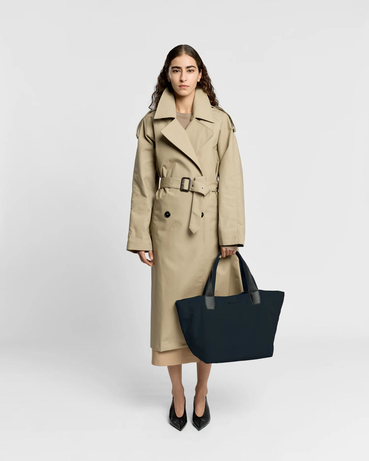 Woman in a beige trench coat holding the Porter Essence Medium Tote Bag in Midnight Trench by Vee Collective against a plain studio background.