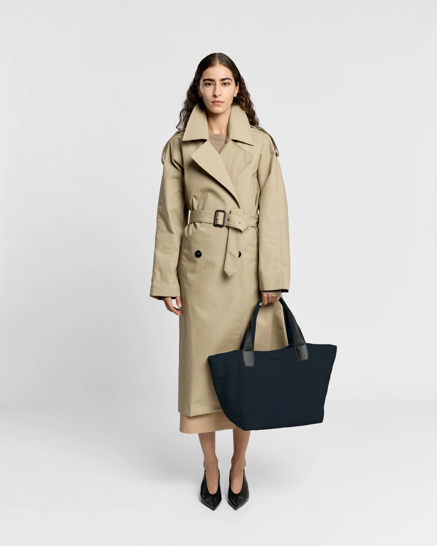 Woman in a beige trench coat holding the Porter Essence Medium Tote Bag in Midnight Trench by Vee Collective against a plain studio background.