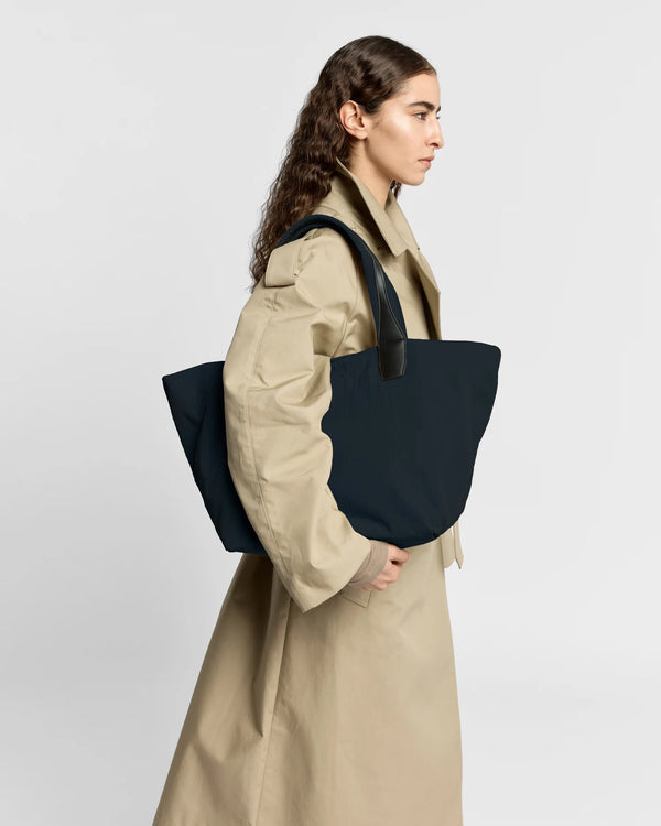 Woman in a beige trench coat carrying the Porter Essence Medium Tote Bag in Midnight Trench by Vee Collective against a plain studio background.