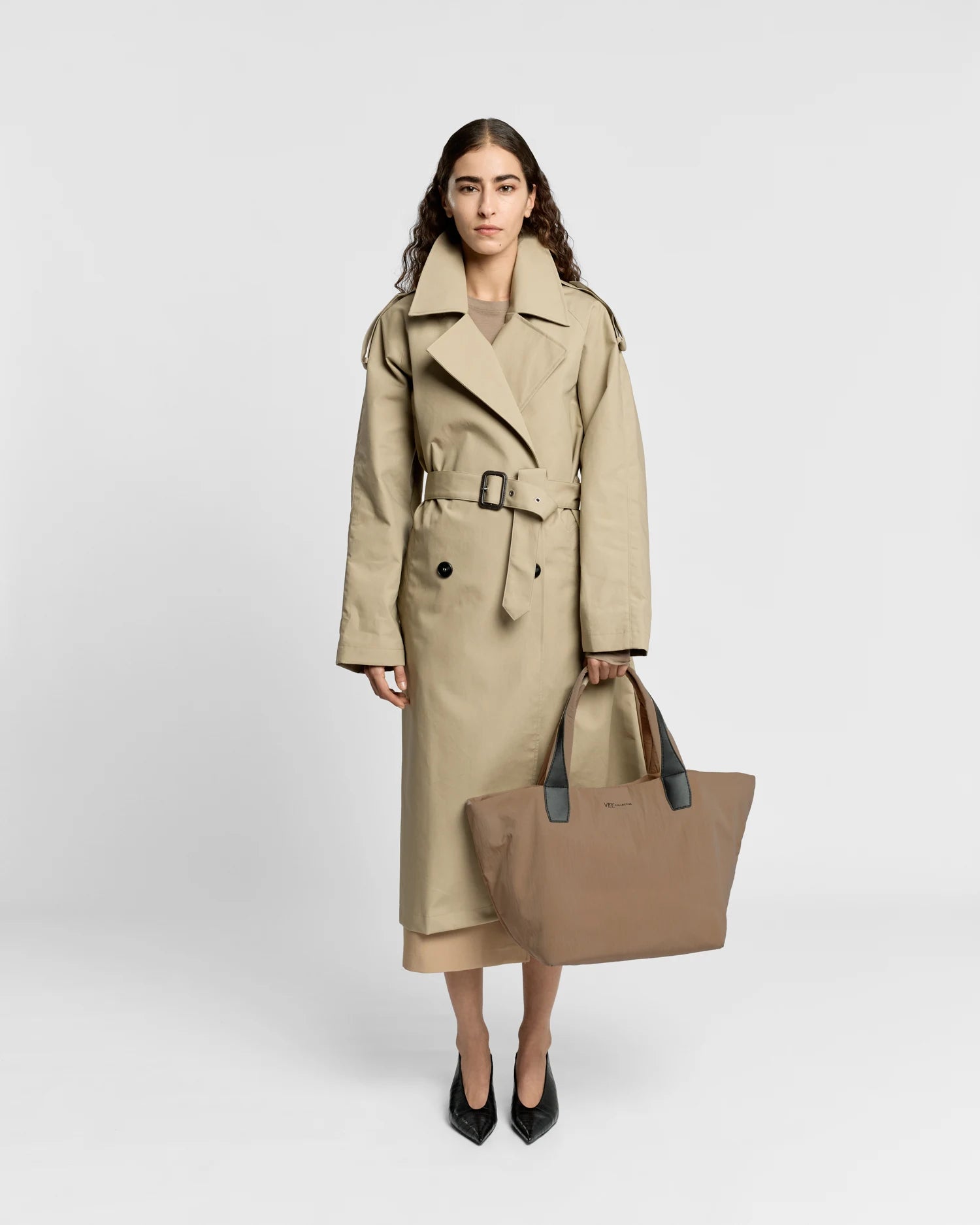 Woman in a beige trench coat holding the Porter Essence Medium Tote Bag in Mink Trench by Vee Collective against a plain studio background.