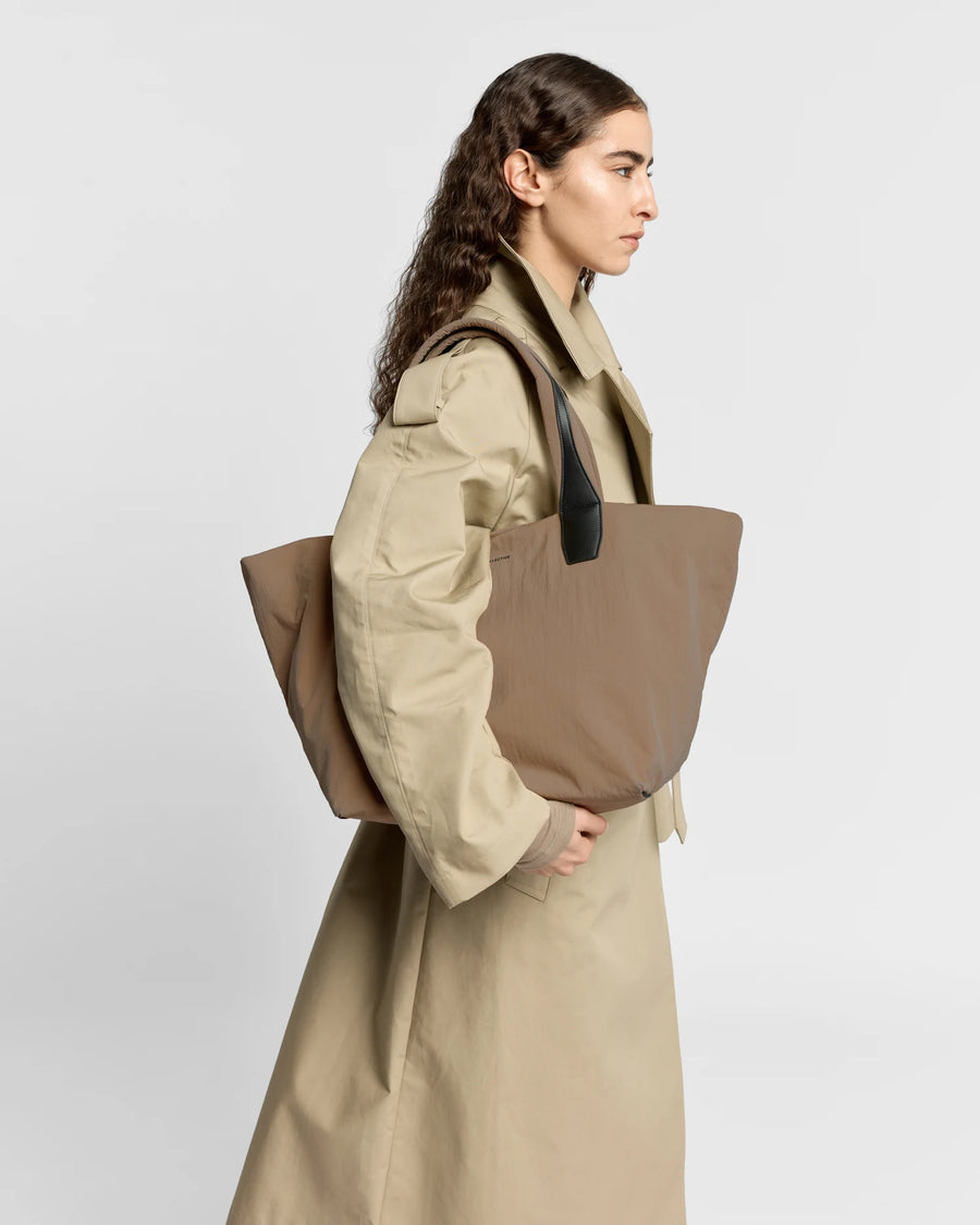 Woman in a beige trench coat carrying the Porter Essence Medium Tote Bag in Mink Trench by Vee Collective against a plain studio background.
