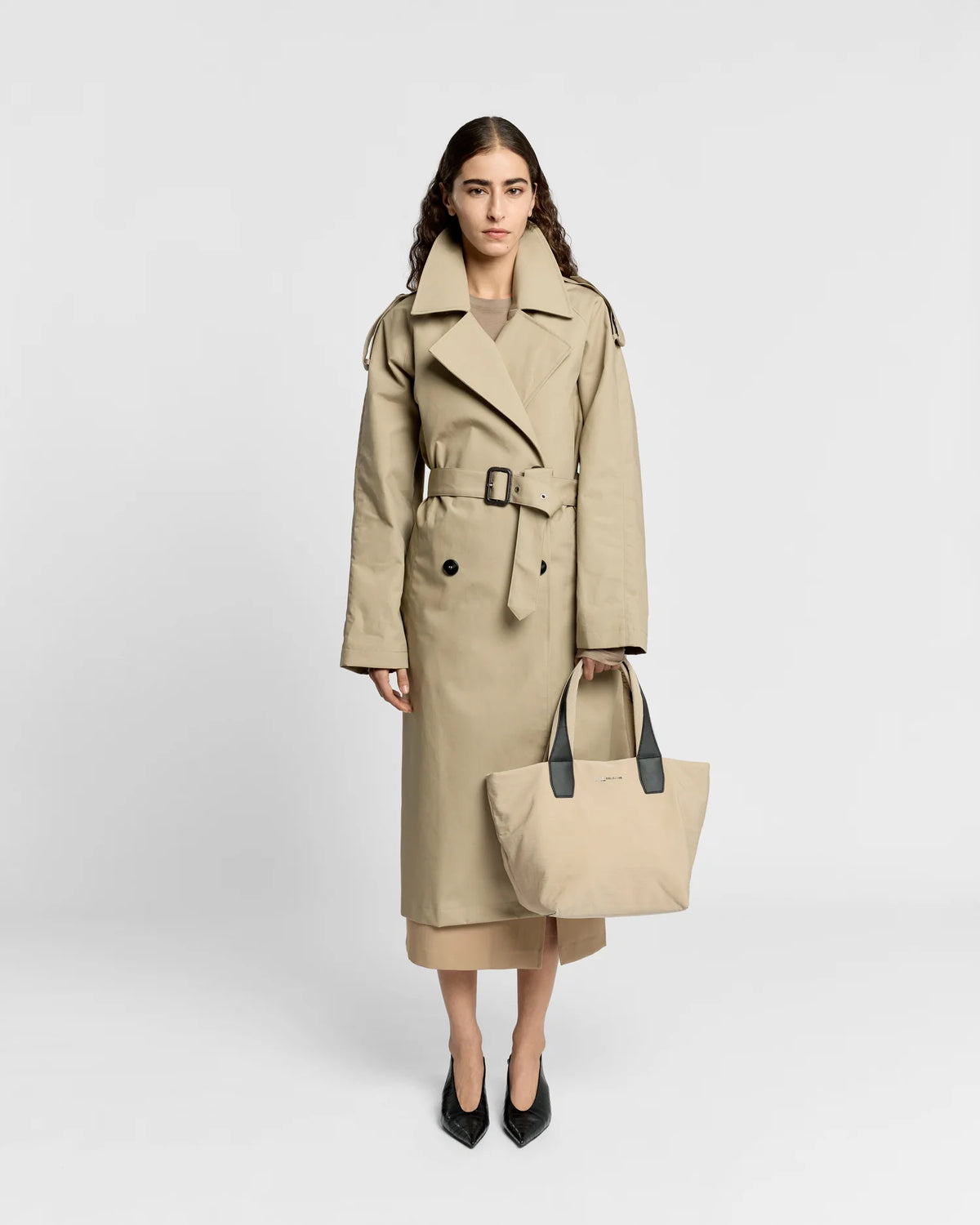 Woman in a beige trench coat holding the Porter Essence Small Tote Bag in Beige Trench by Vee Collective against a plain studio background.