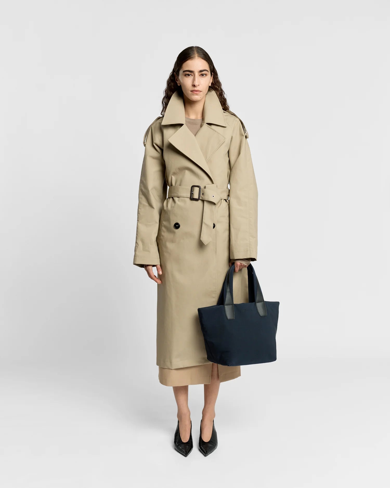 Woman in a beige trench coat holding the Porter Essence Small Tote Bag in Midnight Trench by Vee Collective against a plain studio background.