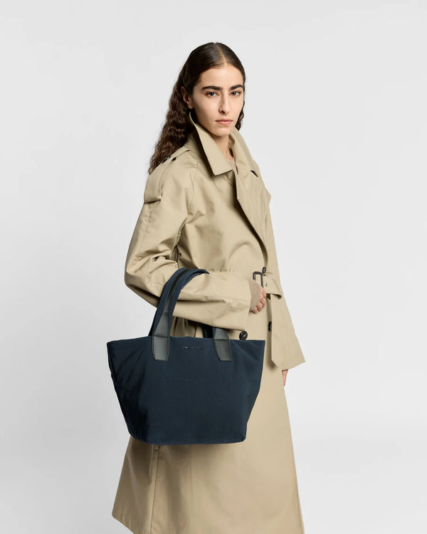 Woman in a beige trench coat carrying the Porter Essence Small Tote Bag in Midnight Trench by Vee Collective against a plain studio background.