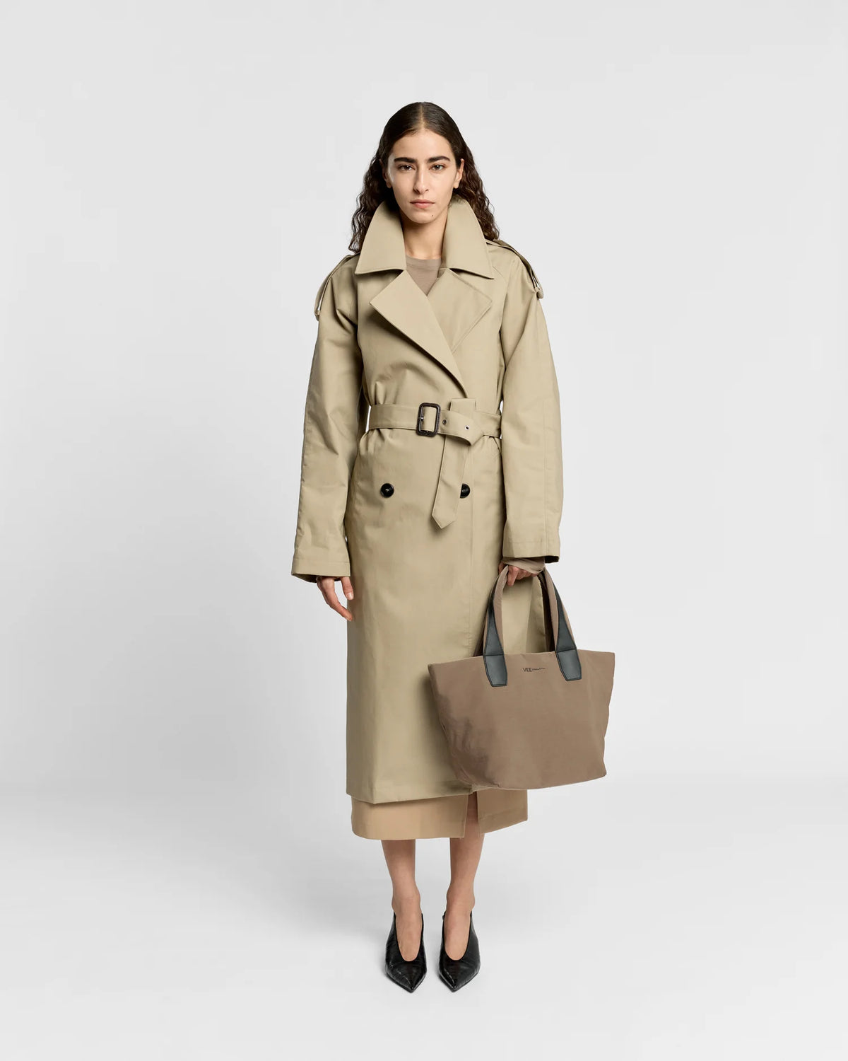 Woman in a beige trench coat holding the Porter Essence Small Tote Bag in Mink Trench by Vee Collective against a plain studio background.